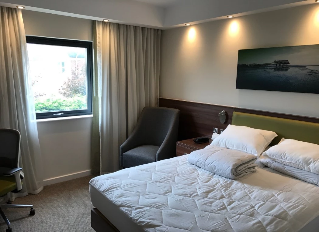 A neatly made bed with pillows and folded linens sits in a contemporary hotel room, featuring a window with curtains, a grey armchair, a desk chair, and a wall painting.
