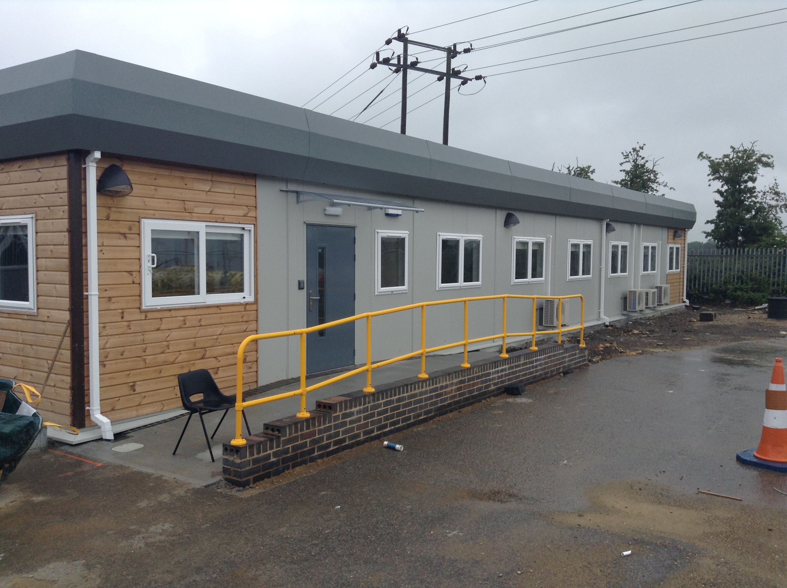Rectangular modular building with teal and gray panels; situated on a newly prepared plot encircled by a construction path. Leafless trees and residential homes are visible in the background.