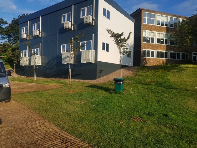 A black, modern three-story building with multiple rectangular windows and a large vertical silver exhaust pipe on the left side; situated outdoors with trees visible in the background.