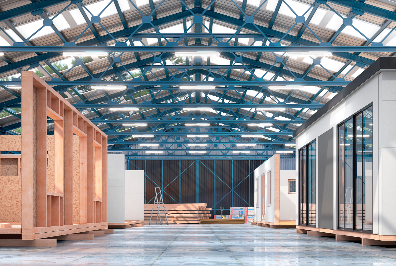 Unfinished modular homes inside a spacious warehouse showcase wooden frames and windows amidst stacks of materials and construction ladders, beneath a high, blue-steel truss roof with skylights.