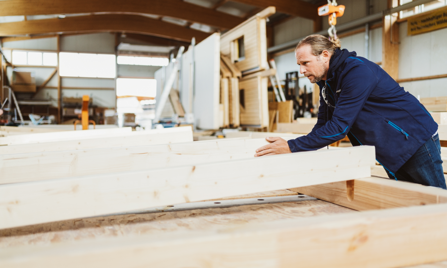 A person wearing a blue jacket inspects wooden planks in a well-lit workshop, surrounded by construction materials and tools.