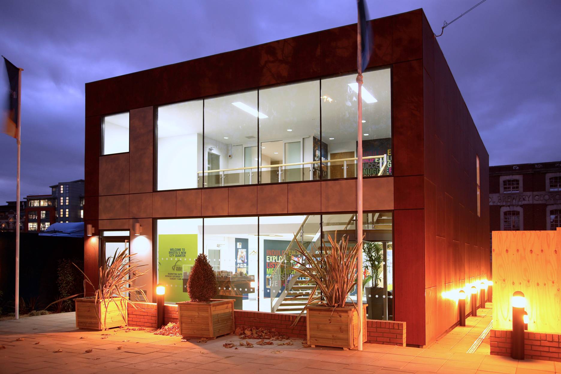 Modern, two-story building with large windows; indoor lights illuminate the interior, featuring stairs and potted plants; exterior includes planters and pathway lights; surrounded by a twilight urban environment. Text: “Welcome to York House” and other smaller print.