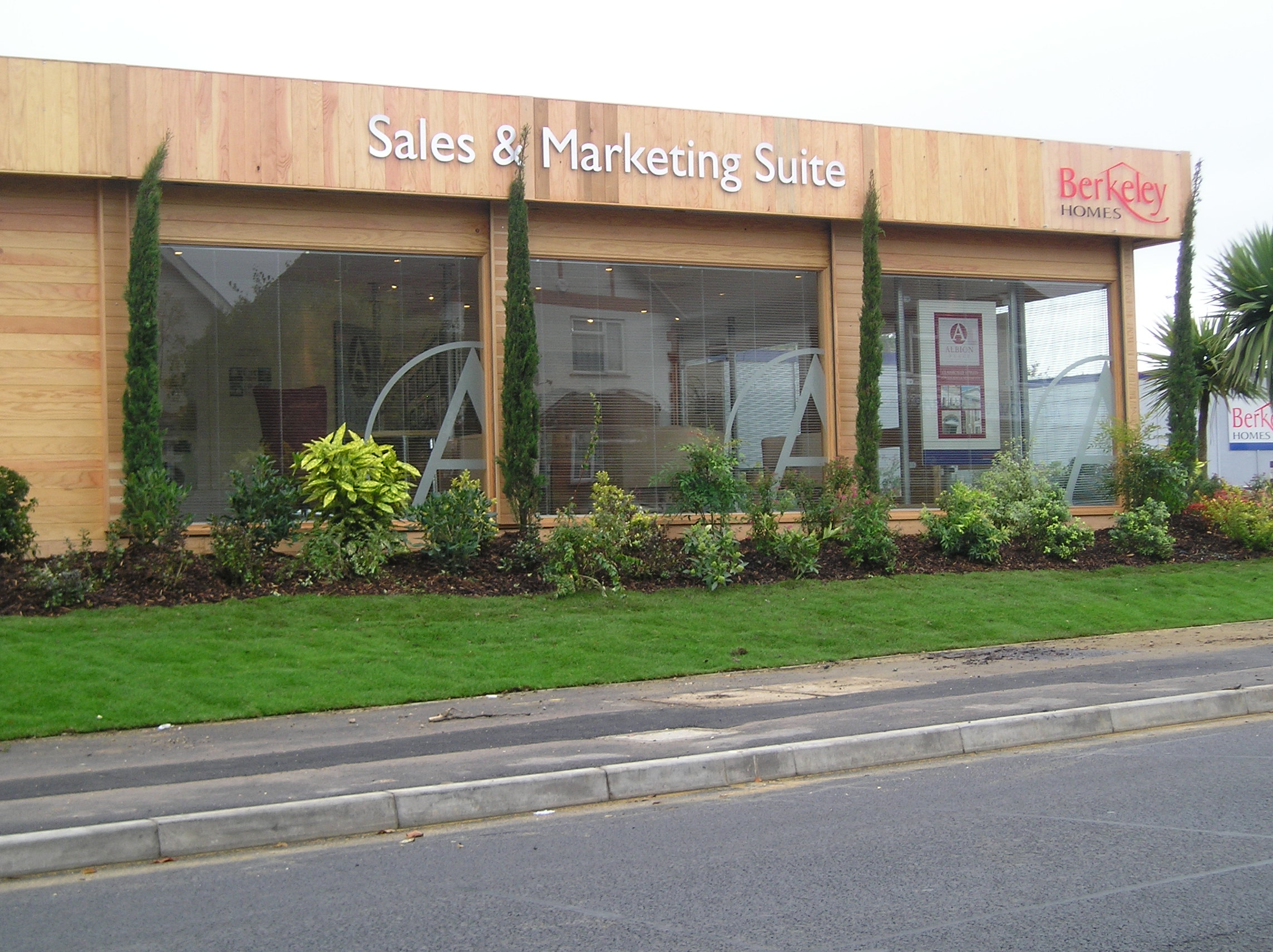 A wooden building labeled "Sales & Marketing Suite" with a "Berkeley Homes" sign is surrounded by greenery, including bushes and tall trees, located adjacent to a paved road and sidewalk.