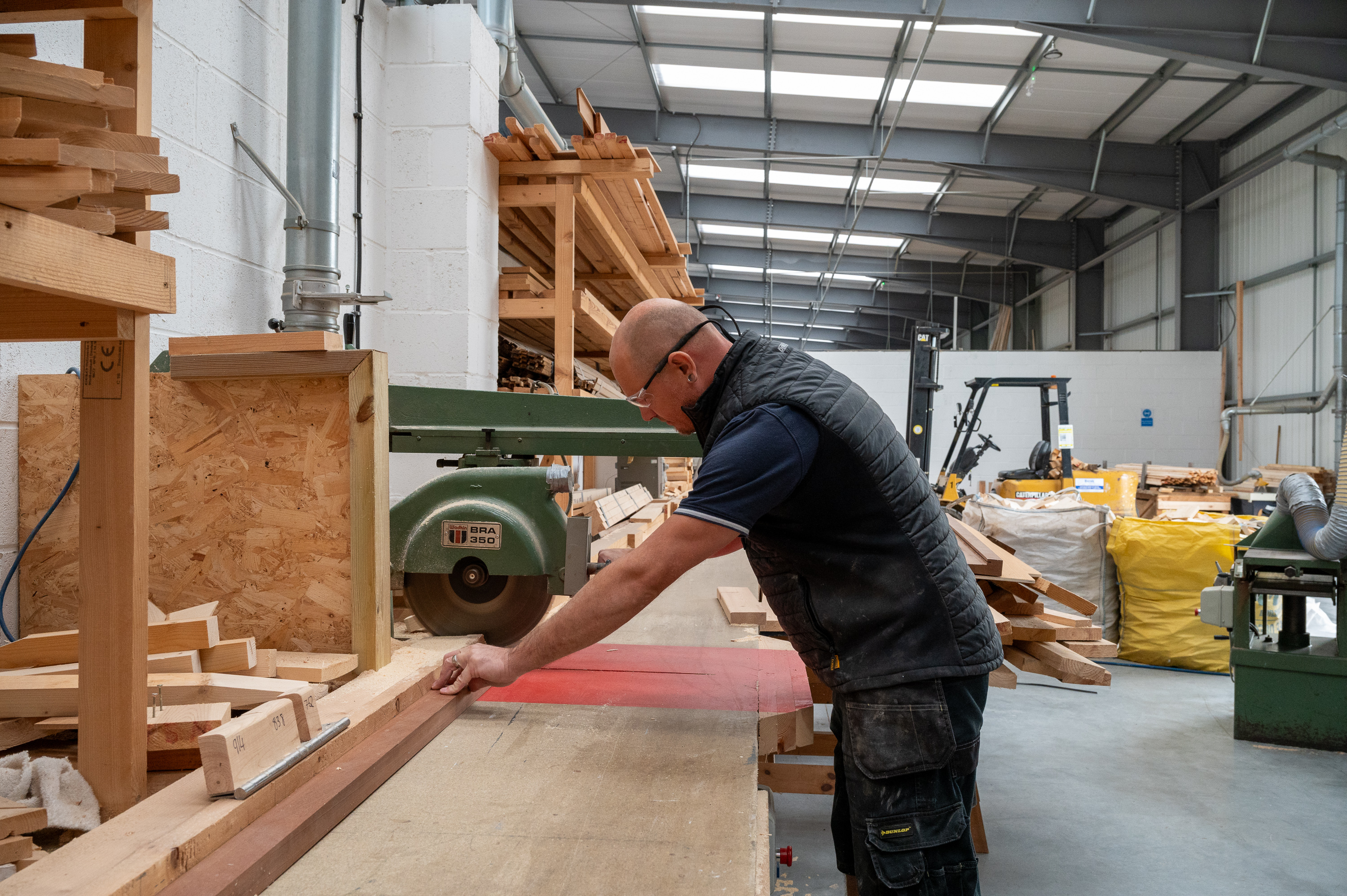 A man operating a woodcutting machine indoors, surrounded by wooden planks and carpentry tools in a spacious, well-lit workshop.