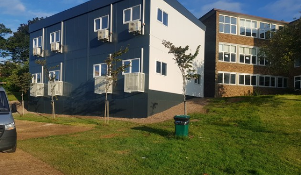 A two-story building with white and gray facade, equipped with air conditioning units, is situated on a grassy hill near a van and trees, adjacent to another brick building.
