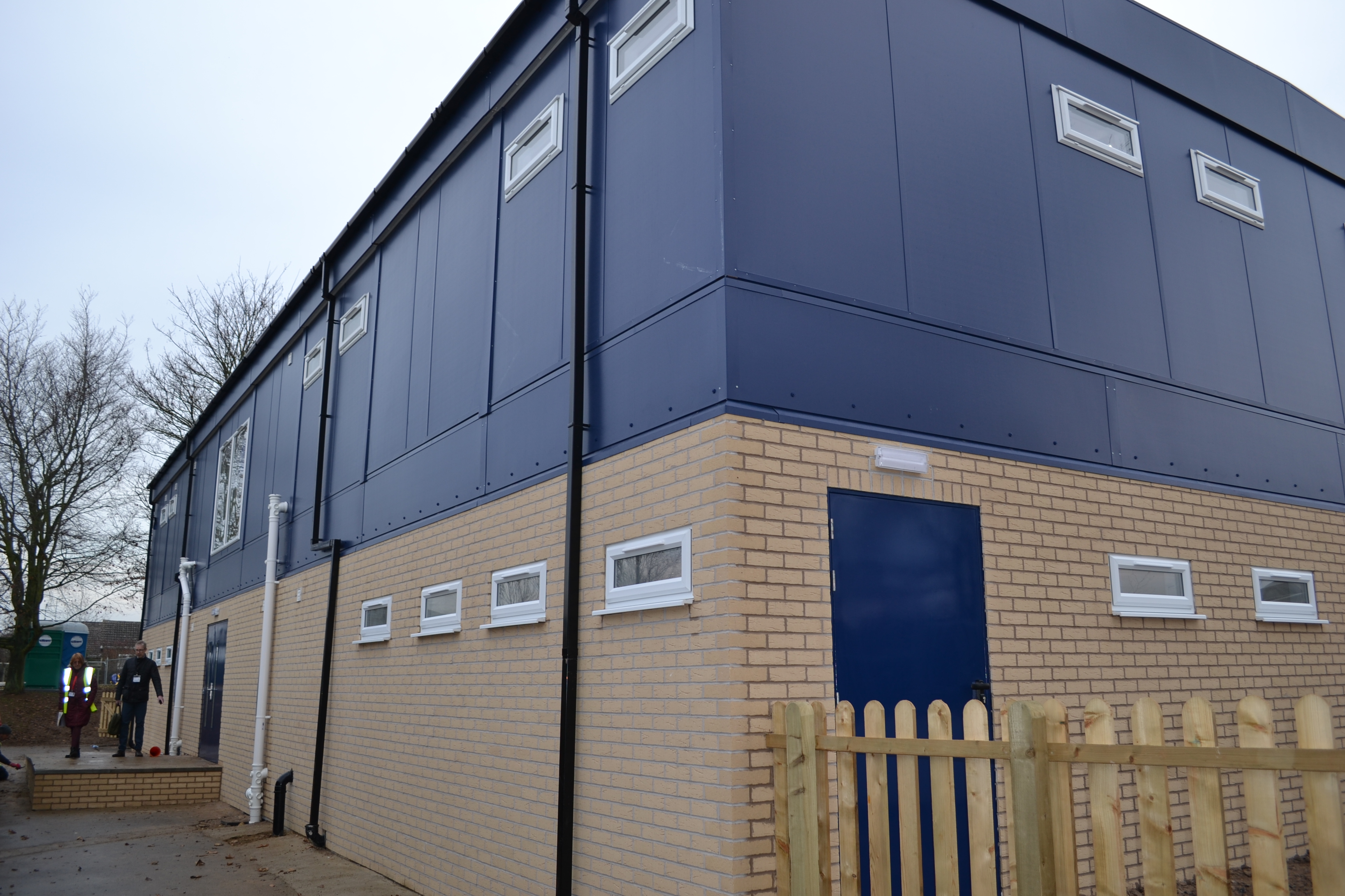 A two-story building with beige brick on the lower level and blue paneling on the upper, featuring several windows. Two people stand nearby; bare trees are visible in the background.