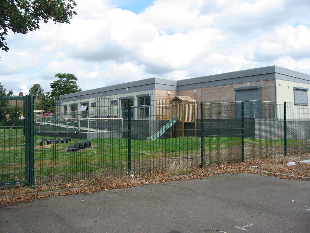 A modern, single-story building with large windows and an adjacent jungle gym stands behind a tall, metal fence in a grassy area under a partly cloudy sky.
