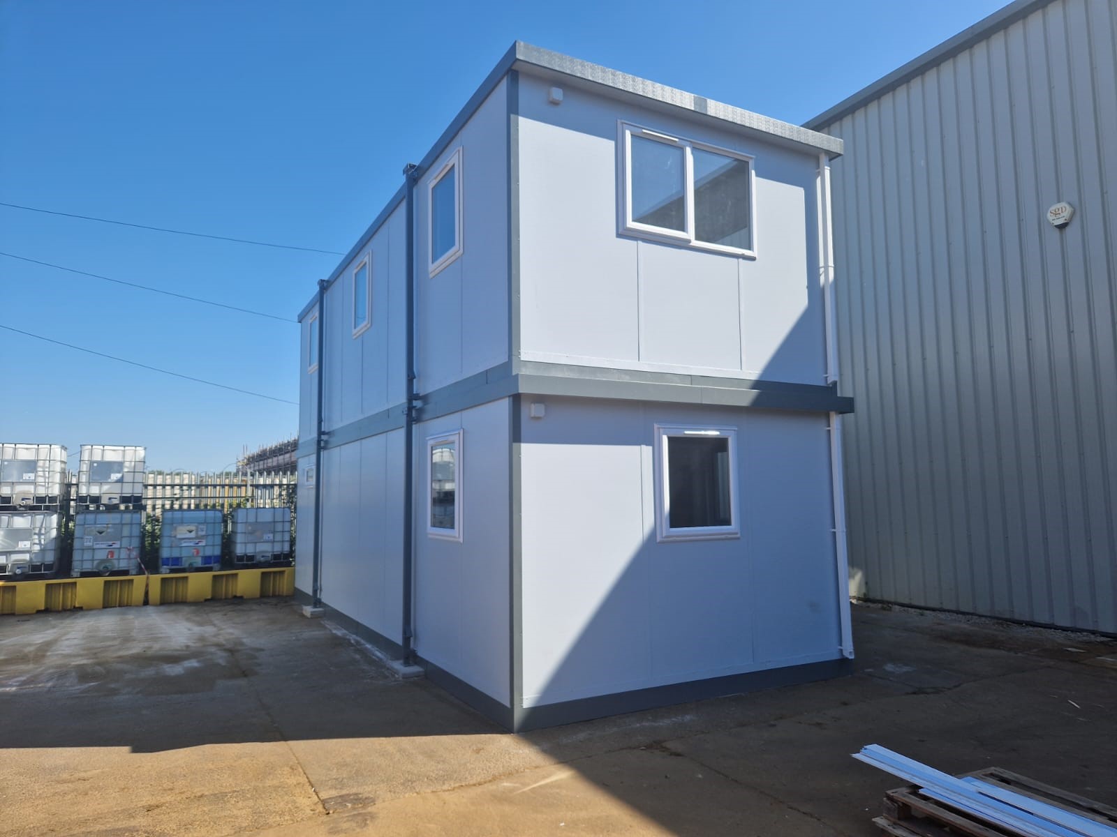 Two stacked portable buildings sit on a concrete lot, adjacent to a metal warehouse. Stacked containers are in the background, and discarded materials lay near the lower right corner.