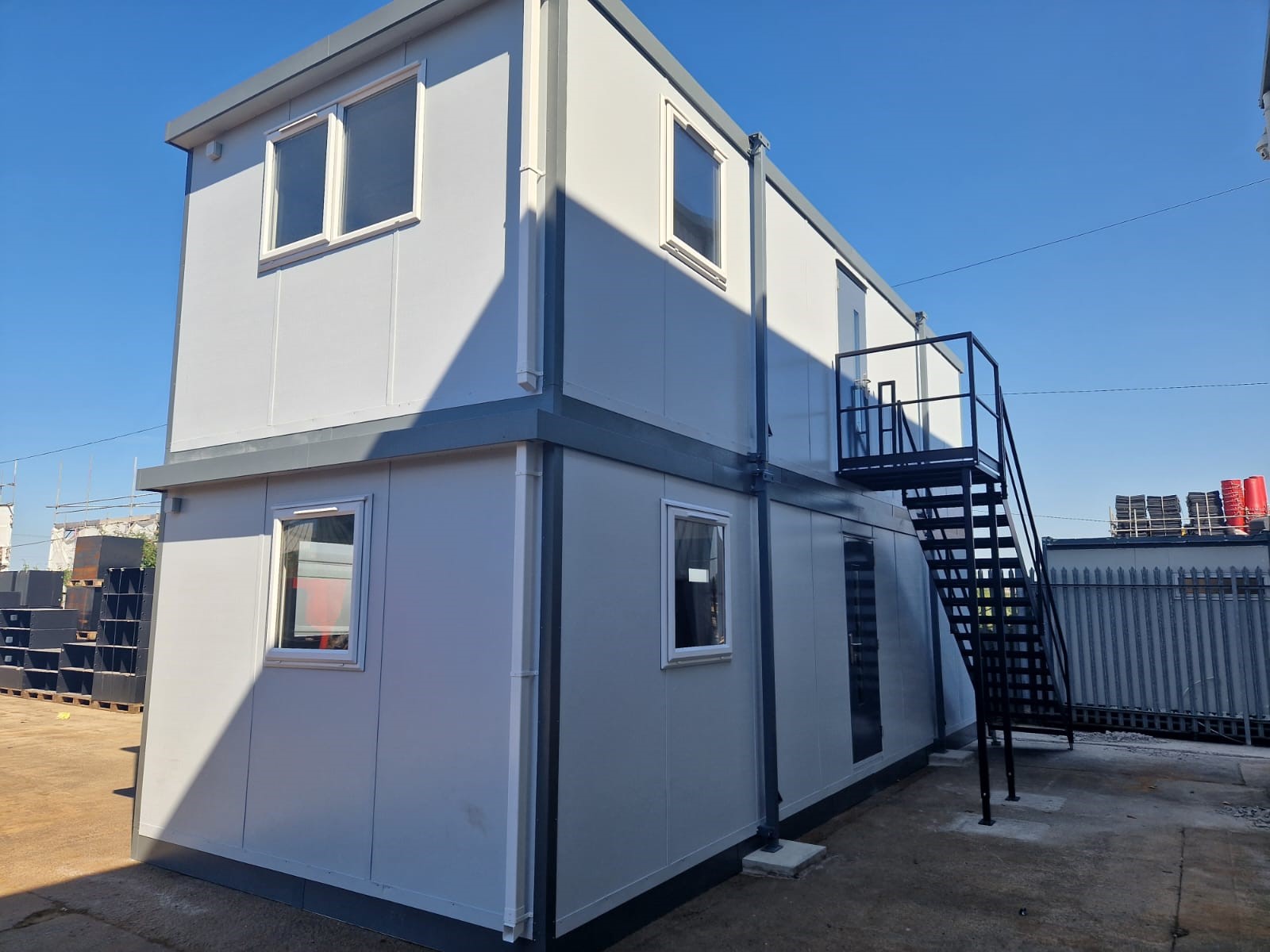 A two-story portable building stands on a concrete yard with external stairs leading to the upper floor, surrounded by industrial materials and a metal fence under a clear blue sky.